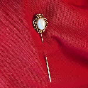 Op de afbeelding: Een gouden broche met een witte ovale steen in een decoratief frame. De broche is bevestigd aan een dunne gouden pin.