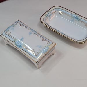 Pode incluir: Conjunto de porcelana de duas peças com uma caixa retangular para bugigangas e uma bandeja a condizer. Ambas as peças apresentam uma base branca com detalhes florais azuis, acabamento dourado e uma borda azul clara. A caixa tem tampa e pés.