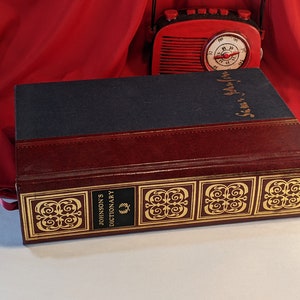 May include: A burgundy leather-bound book with gold accents and the text "Johnson's Dictionary" on the spine. The book is closed and sitting on a white surface.