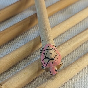 Pode incluir: Um conjunto de espetos de madeira com desenhos de tartarugas pintadas de rosa e preto. Os espetos estão dispostos em forma de cruz.