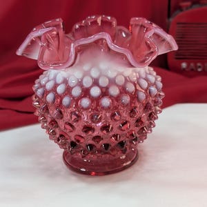 Puede incluir: Un jarrón de cristal rosa con diseño hobnail y borde con volantes. El jarrón tiene un cuerpo redondo con una superficie texturizada de puntos en relieve. El borde es de un tono rosa más claro, y la base es rosa más oscuro.