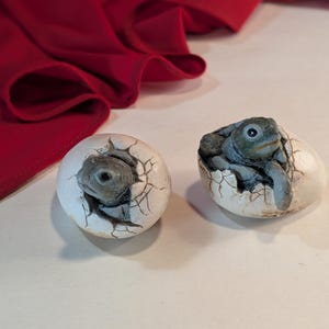 Może przedstawiać: Dwie małe figurki żółwi wykluwają się z białych skorup jaj. Żółwie mają szare pancerze i patrzą z jaj.