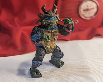 Pode incluir: Uma figura de ação das Tartarugas Ninja Mutantes Adolescentes, de cor preta e verde, com um capacete e armadura de samurai azuis e dourados. A figura está de pé com os braços levantados em uma pose de luta.