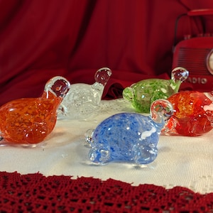 May include: Five glass turtle figurines in various colours, including orange, blue, green, and clear. The turtles are sitting on a white lace tablecloth.