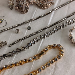 May include: A set of four rhinestone jewellery pieces, including a gold chain bracelet, a silver chain bracelet, a silver chain necklace, and a silver bow brooch. The jewellery is all adorned with clear rhinestones.