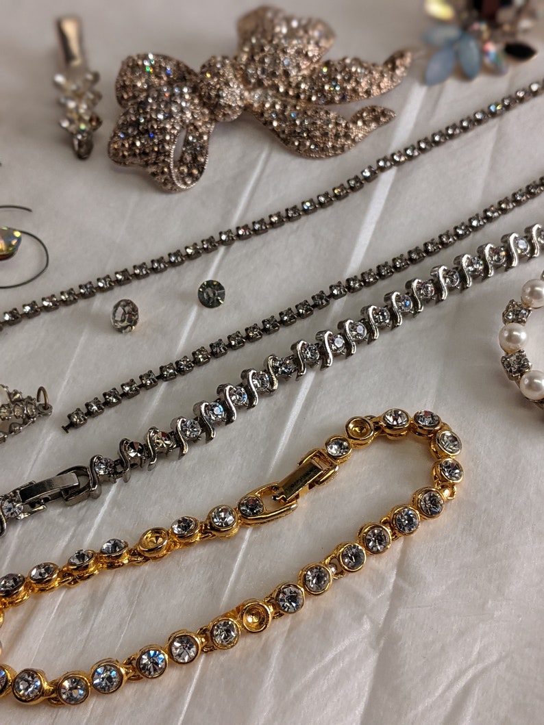May include: A set of four rhinestone jewellery pieces, including a gold chain bracelet, a silver chain bracelet, a silver chain necklace, and a silver bow brooch. The jewellery is all adorned with clear rhinestones.