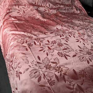 Puede incluir: Un cubrecama de satén rosa con un patrón floral. El patrón está en relieve y presenta un diseño repetitivo de hojas y flores.