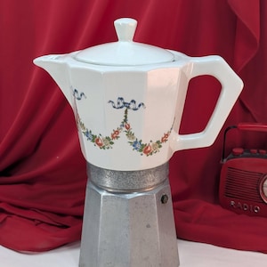 Puede incluir: Una cafetera italiana de cerámica blanca con un diseño floral y una base plateada. La cafetera tiene un asa y una tapa. El diseño floral incluye lazos azules y flores coloridas. Una radio roja está en el fondo.