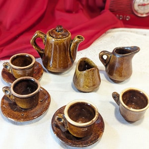 May include: A brown ceramic tea set with a teapot, creamer, sugar bowl, and four cups and saucers. The set is arranged on a white tablecloth.