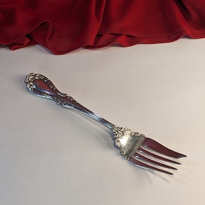 Puede incluir: Un tenedor de plata con un mango adornado y tres dientes. El tenedor está sobre una superficie blanca.