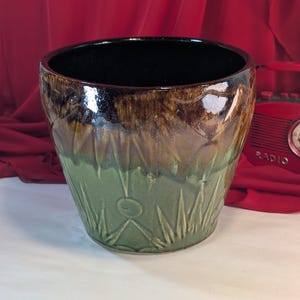 May include: A ceramic planter with a glossy finish. The top is dark brown, transitioning to a lighter brown and then to a green base with embossed leaf and circle designs. The interior is black.