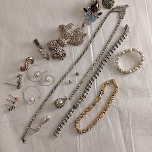 May include: A collection of vintage jewellery pieces, including a rhinestone necklace, a bracelet, a ring, earrings, and brooches. The jewellery is arranged on a white fabric background.