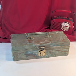 Puede incluir: Una caja de aparejos de pesca de plástico verde con un cierre dorado y un patrón de mármol. La caja tiene una manija de transporte.