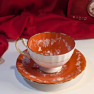 May include: A white teacup and saucer with a floral pattern in white on a bright orange background. The cup and saucer have a gold trim.