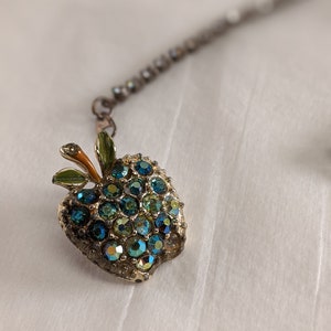 May include: A gold-toned apple charm with green leaves and blue and green rhinestones. The charm is attached to a silver chain.