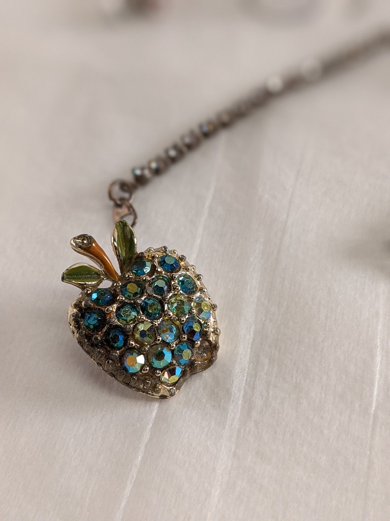 May include: A gold-toned apple charm with green leaves and blue and green rhinestones. The charm is attached to a silver chain.