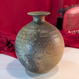 Puede incluir: Un pequeño jarrón redondo de cerámica marrón con un esmalte moteado. El jarrón tiene un cuello estrecho y un labio ligeramente acampanado.