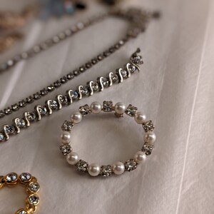 May include: A silver and white rhinestone and pearl ring brooch. The brooch is circular and has a delicate design.