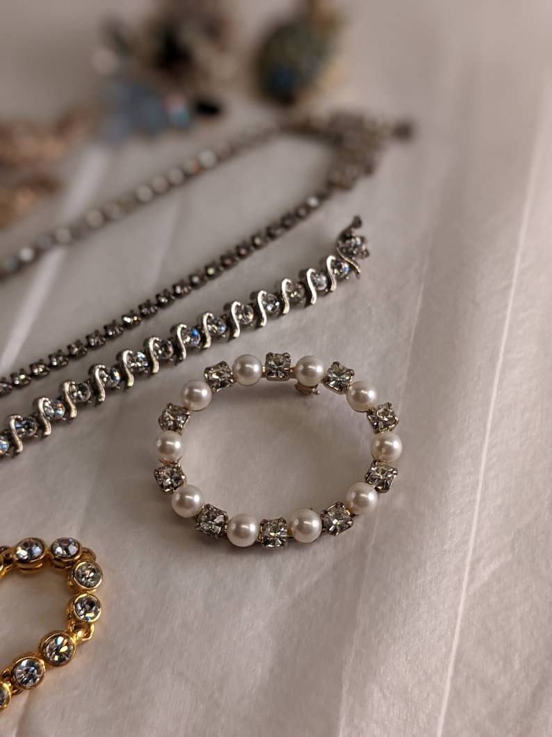 May include: A silver and white rhinestone and pearl ring brooch. The brooch is circular and has a delicate design.