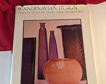Könnte beinhalten: Ein Buch mit dem Titel "Scandinavian Design: Objects of a Life Style" von Eilene Harrison Beer. Der Umschlag zeigt ein Foto einer Sammlung von Glasvasen und einer Schale in verschiedenen Violett- und Orangetönen.