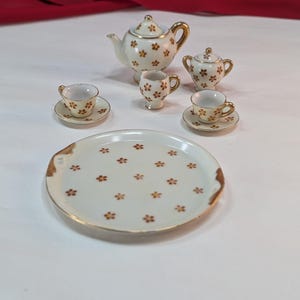 May include: A miniature tea set with a white and gold floral design. The set includes a teapot, sugar bowl, creamer, two teacups with saucers, and a serving tray. The tray is approximately 6 inches in diameter.