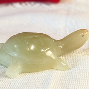 May include: A small, carved turtle figurine in a pale, translucent green colour. The turtle has a detailed shell pattern and is posed with its head and legs extended. The figurine is made of a smooth, polished material.