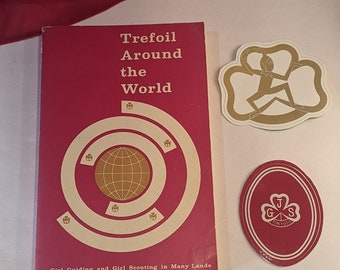May include: A red book titled "Trefoil Around the World" with a white circle and a globe inside. The book is about Girl Guiding and Girl Scouting in Many Lands. There are two patches, one with a gold trefoil and a figure in the centre and the other with a gold trefoil and the letters G, J, and S in the centre.