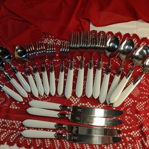 May include: A set of cutlery with white handles. The set includes spoons, forks, and knives. The cutlery is arranged on a red and white lace tablecloth.
