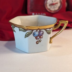 Könnte beinhalten: Eine weiße Porzellan-Teetasse mit goldfarbenem Rand und einem floralen Design mit blauen und grünen Beeren. Die Tasse hat eine sechseckige Form und einen goldenen Henkel.