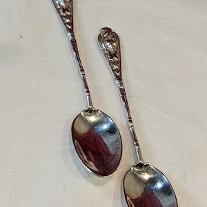 May include: Two silver teaspoons with ornate handles and oval bowls. The spoons are on a white surface.