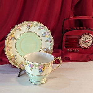 May include: A vintage teacup and saucer set with a delicate floral design. The cup and saucer are both pale green with gold trim and feature a pattern of pink, yellow, and blue flowers. The saucer has a scalloped edge.