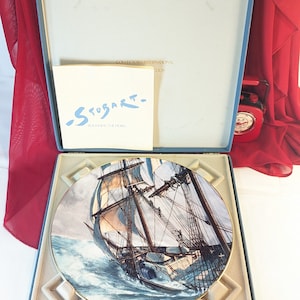 Pode incluir: Um prato de porcelana azul e branco com borda dourada que apresenta uma pintura de um navio navegando no oceano. O prato está em uma caixa azul com interior branco e o texto "Collectors International Edition" e "Stobart - Rounding the Horn" na caixa.