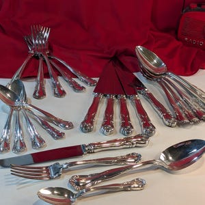 Könnte beinhalten: Ein Besteckset, bestehend aus Gabeln, Löffeln und Messern, mit verzierten Griffen. Das Besteck ist auf einer weißen Oberfläche mit einem roten Tuch im Hintergrund angeordnet.