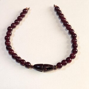 May include: A dark red beaded bracelet with a brown clasp. The bracelet is made of small, round beads and is approximately 18 cm in length.