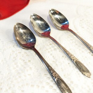 以下が含まれることがあります： 装飾的な花の模様が柄に施された銀色のスプーンが3本、斜めに配置されています。白いレースのテーブルクロスの上に置かれ、左上には赤い布があります。スプーンの長さは約18cmです。