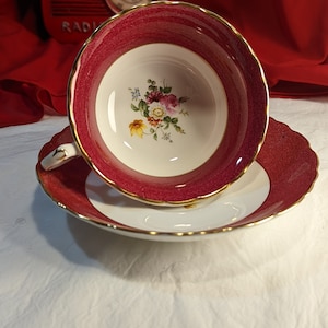 Op de afbeelding: Een vintage theekopje en schotelset met een roze en wit bloemenontwerp. Het theekopje heeft een gegolfde rand en de schotel heeft een gouden rand.