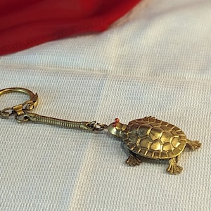 May include: A gold-coloured turtle keychain with red eyes. The turtle is attached to a gold-coloured chain with a key ring.