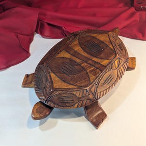 May include: A wooden turtle ornament, intricately carved with a detailed shell pattern. The turtle is a warm brown colour, with lighter wood grain visible. It has four legs and a small tail. A red fabric is in the background.