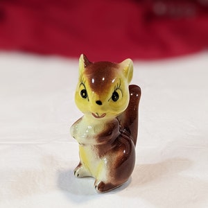 Puede incluir: Una pequeña figurita de ardilla de cerámica pintada en marrón y amarillo. La ardilla tiene una cola marrón y está de pie sobre sus patas traseras.