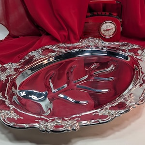 May include: A silver oval serving platter with decorative floral edging. The platter features a central, stylized design. The background includes red fabric and a vintage radio.