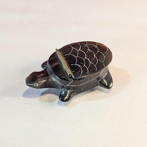 May include: A small, dark brown turtle-shaped box with a hinged lid. The box is made of wood and has a carved design on the top. The lid is open and the box is empty.