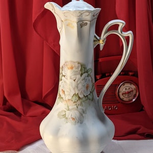 Könnte beinhalten: Eine weiße Porzellan-Kanne mit goldener Verzierung und Blumenmuster. Die Kanne hat einen hohen, schlanken Körper und einen geschwungenen Griff. Die Blumen sind weiße Rosen mit grünen Blättern.