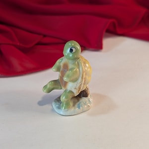 May include: A small ceramic figurine of a green turtle sitting on a log. The turtle has a brown shell and a green head with a single black eye. The figurine is sitting on a white base with a blue and green design.