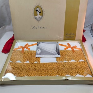 Puede incluir: Un juego de sábanas en caja con un marco dorado. El juego incluye sábanas blancas con ribete de encaje dorado y lazos naranjas. La caja presenta el texto "Lady Christina" y "Luxury Decorative Ensemble".