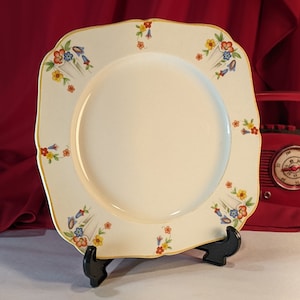 Puede incluir: Un plato cuadrado blanco con un borde dorado y un diseño floral en tonos de azul, rojo, amarillo y naranja.