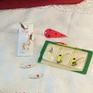 Op de afbeelding: Assortiment van vislokken, waaronder een rood-zwarte lok met zwarte stippen, een zilveren lok met een rode stip en een gele lok met een zwarte stip. De lokken worden gepresenteerd op een wit oppervlak met een rood-witte kantversiering.