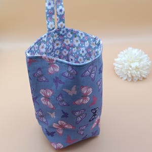 Könnte beinhalten: Ein hängender Aufbewahrungskorb aus Stoff mit einer blauen Außenseite, die mit einem Muster aus rosa, violetten und weißen Schmetterlingen bedruckt ist. Das Innere ist mit weißem Stoff mit kleinen blauen und rosa Blumen gefüttert und hat einen Henkel.