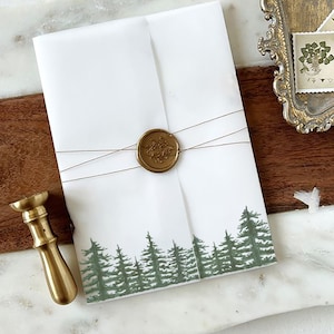 May include: A white envelope with a green forest design at the bottom. The envelope is tied with twine and sealed with a gold wax seal.
