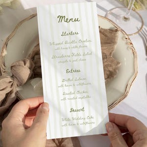 May include: A vertical menu card with a light green and white striped pattern. The word "Menu" is at the top in a script font, followed by the starters, entrees, and dessert options. The menu includes Whipped Ricotta Crostini, Grilled Salmon, and White Wedding Cake.