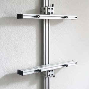 May include: A silver metal shelf unit with two adjustable shelves. The shelves are mounted to a vertical support post with black knobs. The unit is mounted to a white wall.